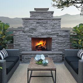 An outdoor living area with a stone fireplace. Two gray loveseats flank the fireplace and there is a coffee table made of wood and metal in between the loveseats.