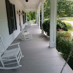 A front porch with white siding and black-trimmed windows is framed by shrubs along the porch. There are three white rocking chairs along the porch and a white porch swing at the end.
